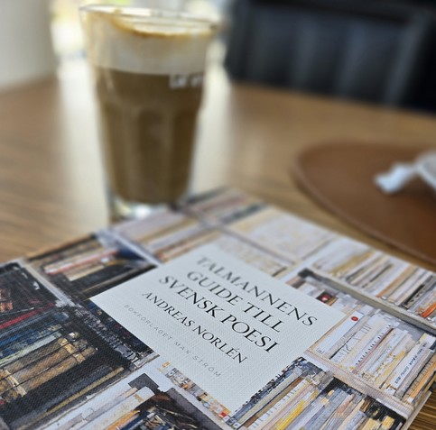 Antologin talmannens guide till svensk poesi på ett träbord framför en caffe latte.